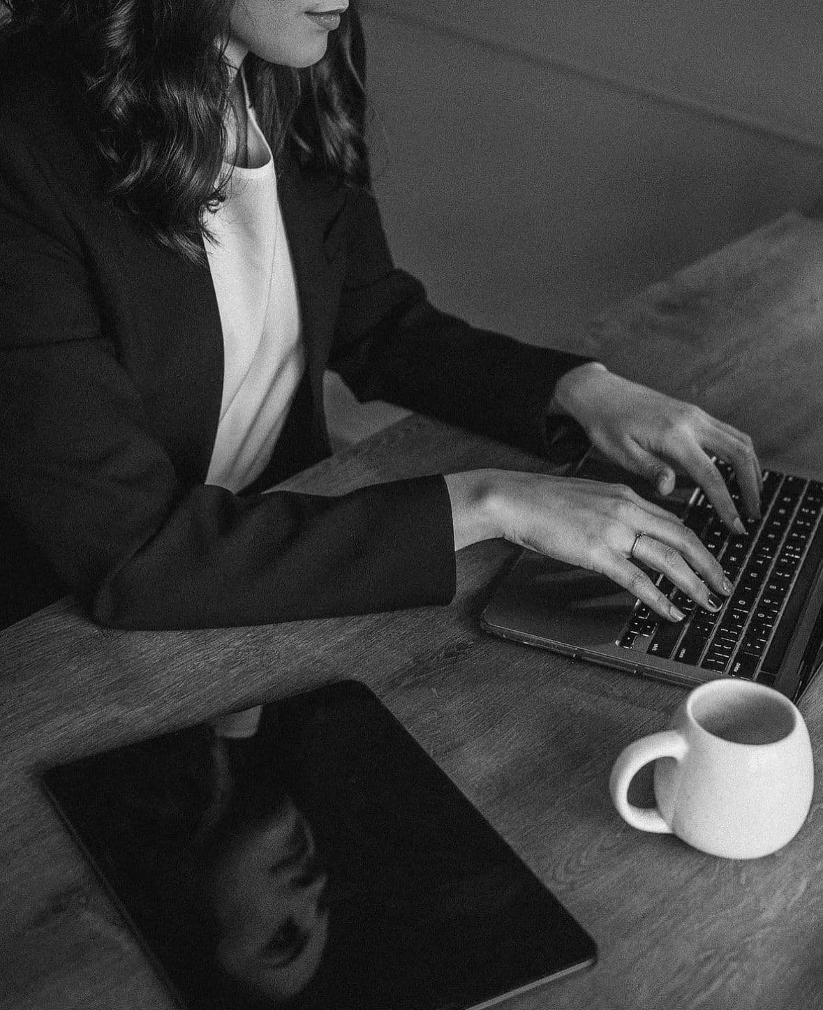 Mulher de blazer digitando no laptop em mesa de madeira com tablet e caneca branca.